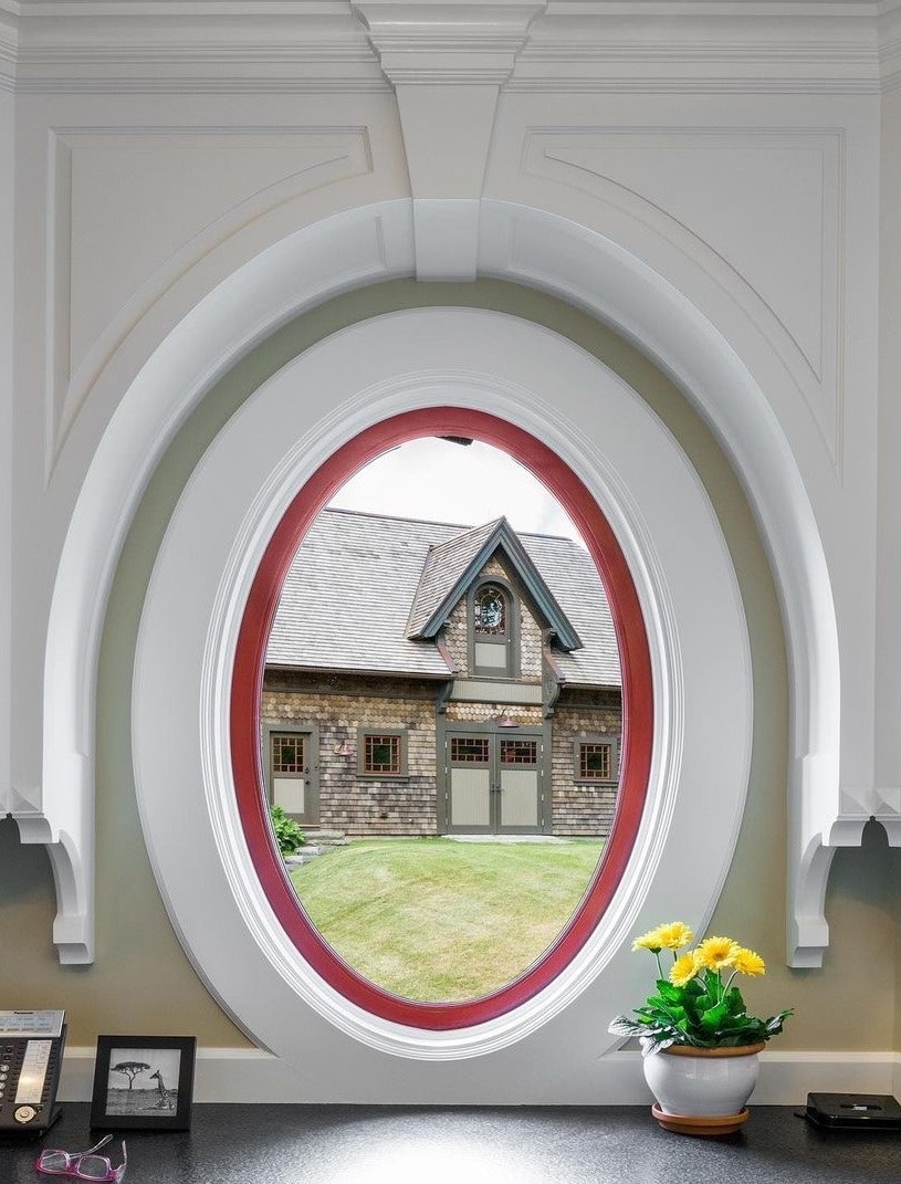A red oval window with white trim sits over a desk and overlooks a house across the yard.