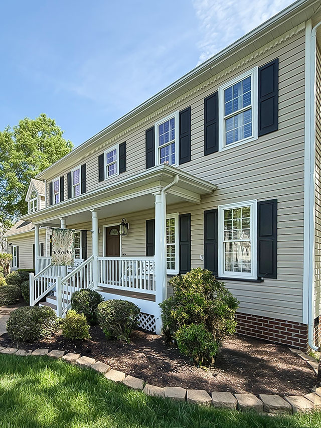 A two-story colonial style house with beige siding, black shutters, and a welcoming front porch. The porch features white railings and columns, a hanging lantern light fixture, and a 'Welcome' sign. The house is surrounded by well-maintained landscaping including bushes and a green lawn.