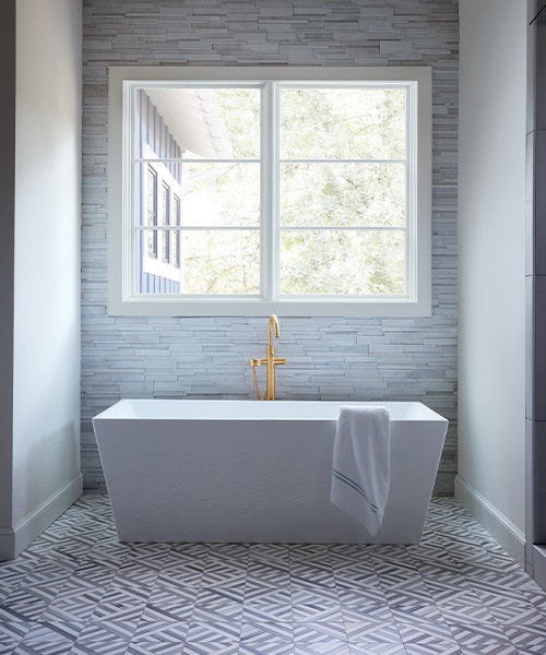 A contemporary soaker tub with gold hardware sits underneath two casement windows