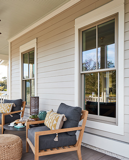Inviting front porch featuring two double-hung windows, wooden chairs with gray cushions and patterned pillows, a wicker ottoman, and decorative accents on a neutral siding backdrop.   