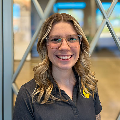 A smiling team member with glasses and tattoos stands in front of a large wall logo.