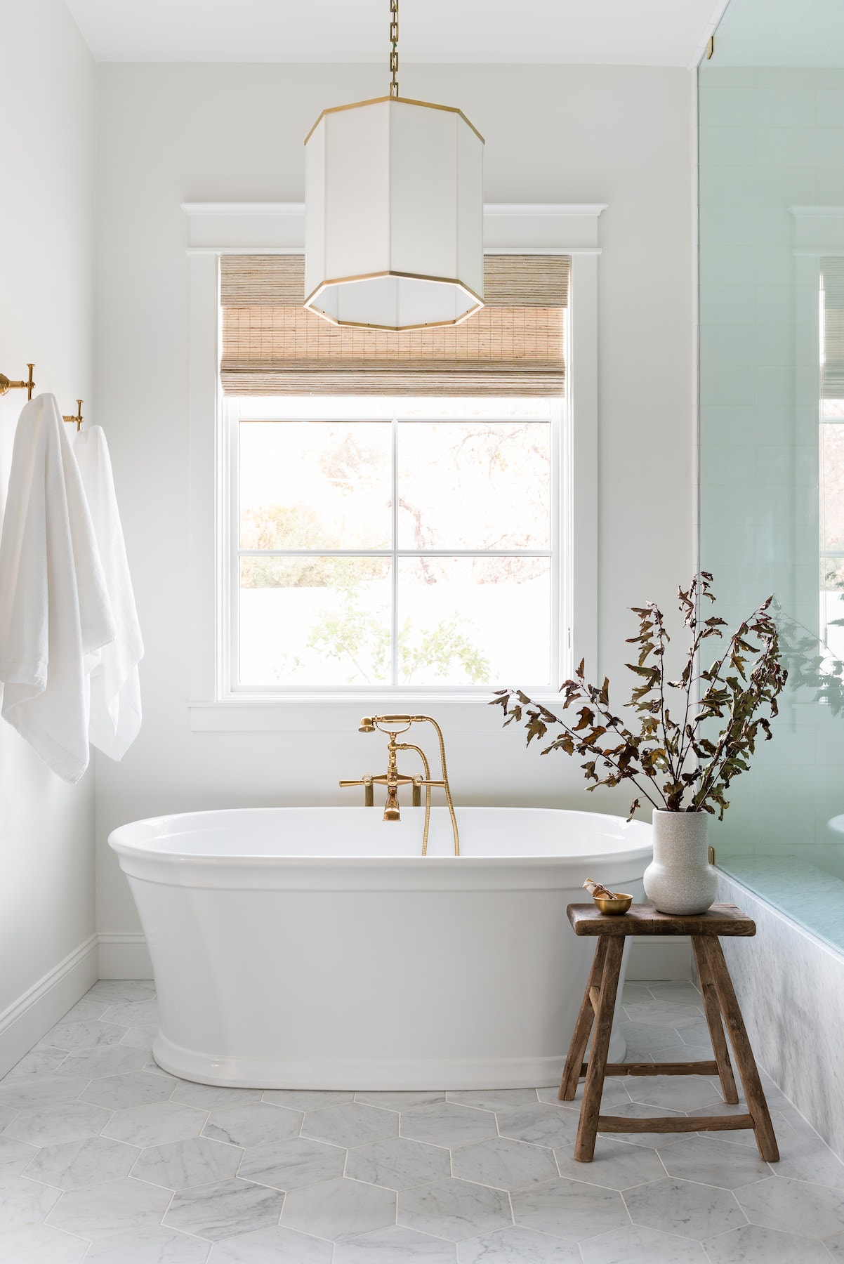 A white picture window with Roman shades sits behind a freestanding bathtub.