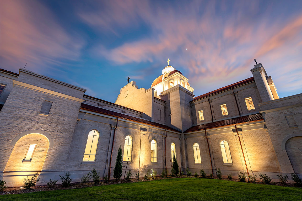 Illuminated church exterior with arched windows and dome under colorful evening sky
