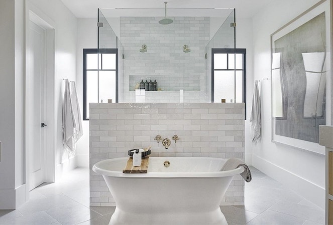 a white soaker tub in front of a white diving wall and two black windows far in the back of the bathroom