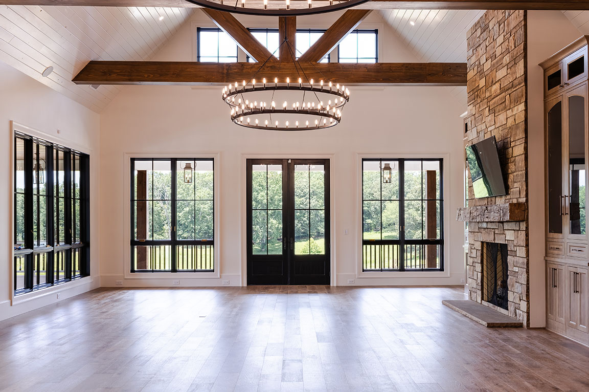 Luxurious great room with a grand chandelier, exposed wooden beams, stone fireplace, and large windows providing natural light.