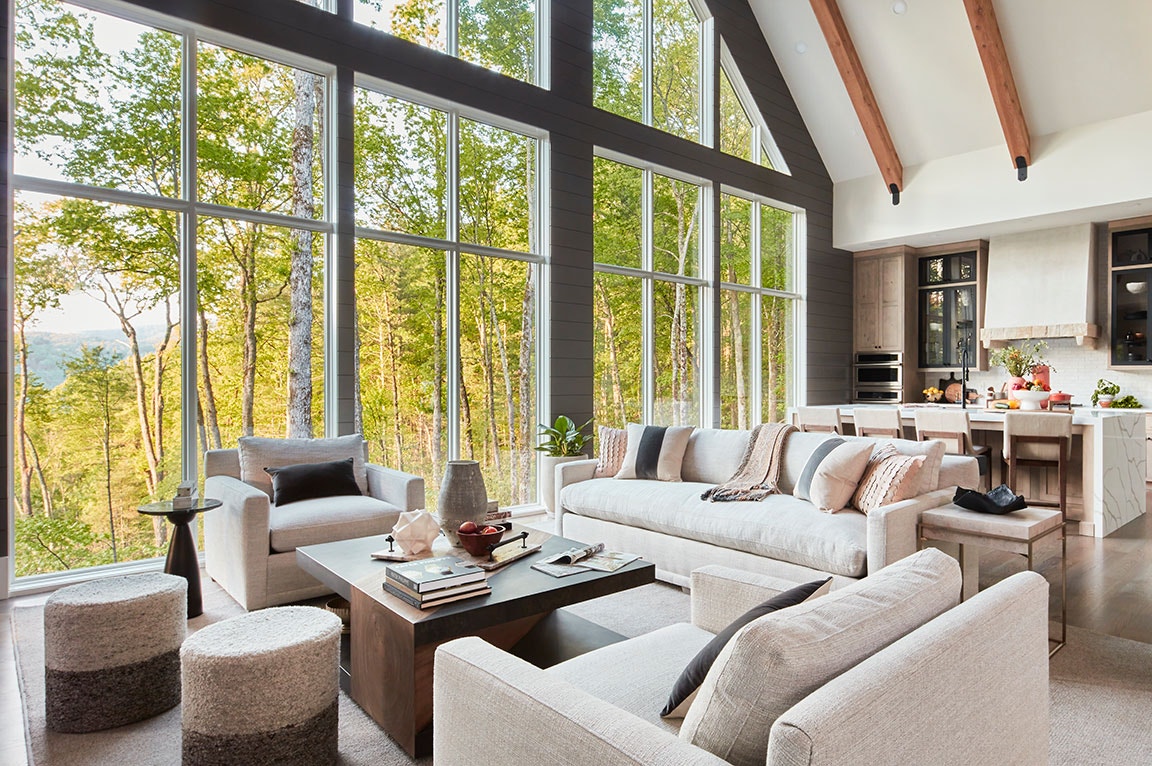 Modern living room with floor-to-ceiling windows, neutral couches, and forest views.