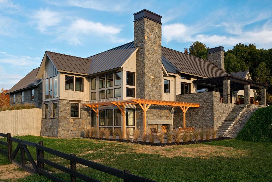 The exterior of a farmhouse-style home has beautiful stonework and basement windows overlooking an outdoor patio.
