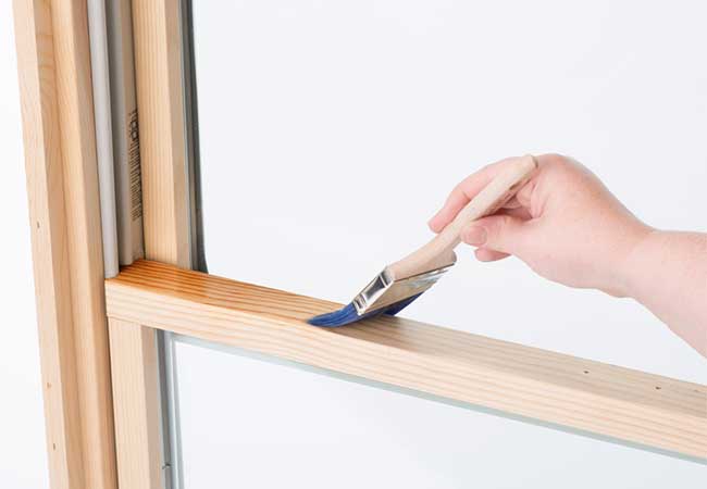 Hand holding a paintbrush applying a first coat of stain to an unfinished wood window.