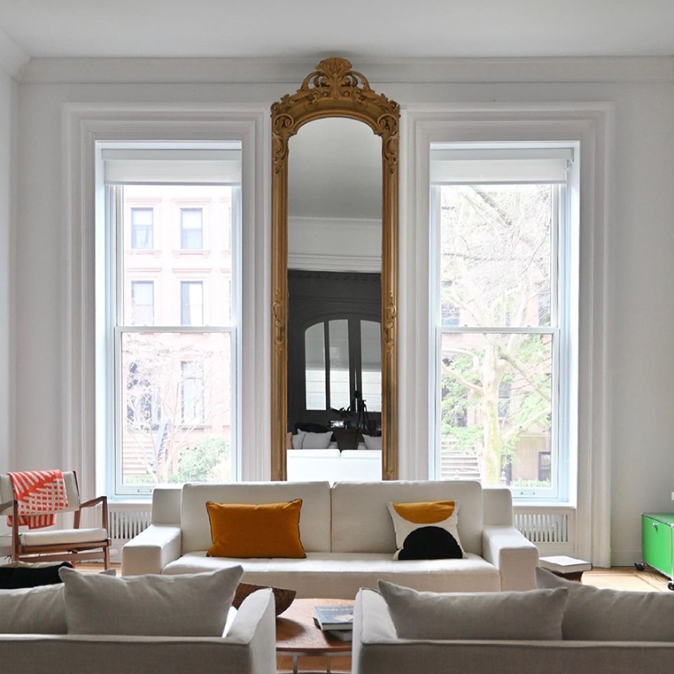 A modern living room featuring brownstone windows, a large mirror, and a cozy sofa with colorful cushions.