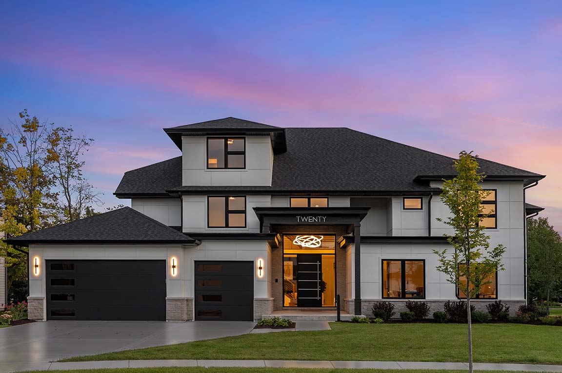 Modern two-story home exterior with dark roof, large windows, and illuminated entry at dusk.
