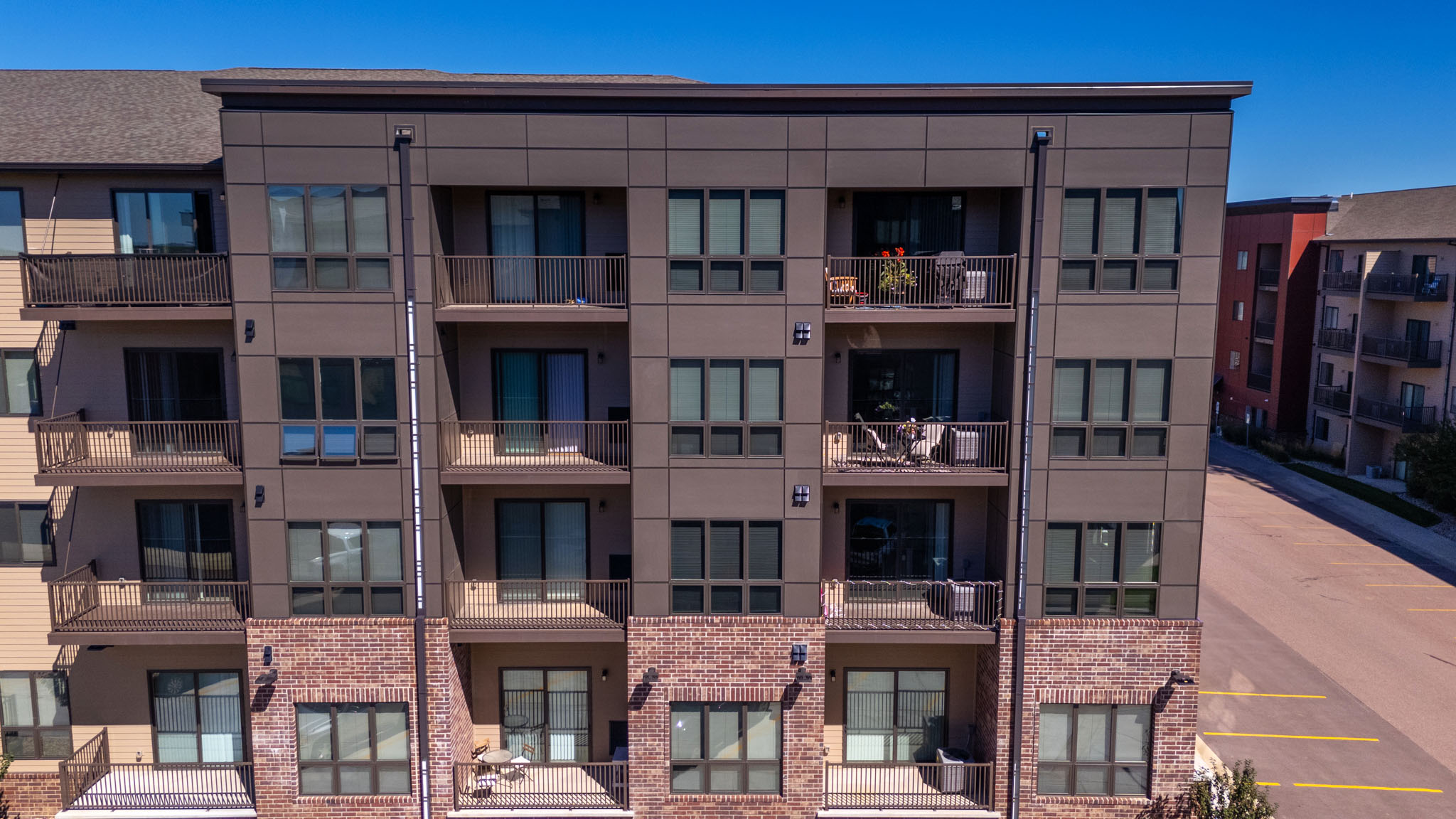 A modern apartment building with large apartment windows and balconies facing a parking lot on a sunny day.