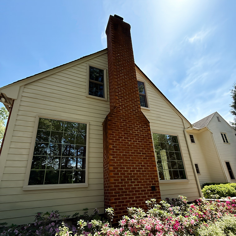 Replacement windows on a white house with a tall brick chimney and blooming bushes in front.