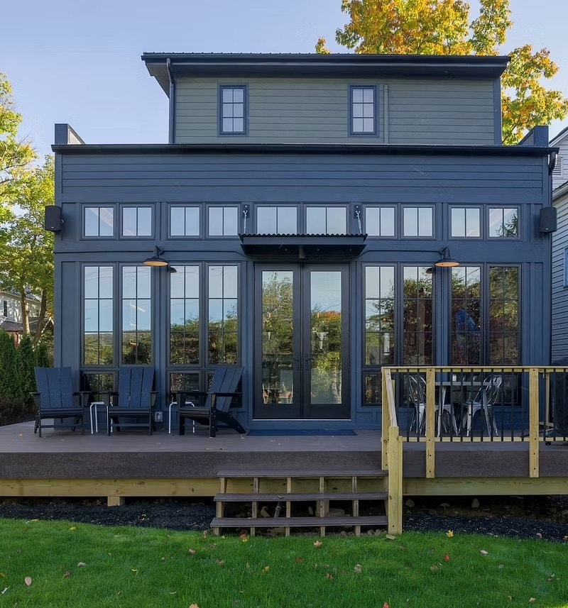 The exterior of a sunroom with black windows, a patio door and siding overlooks a deck.