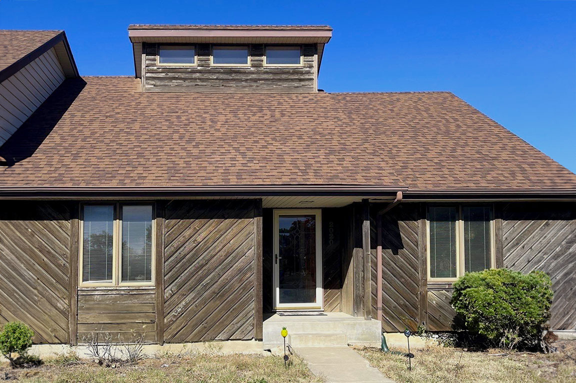A wooden house before window replacement in Topeka, featuring a brown roof and clear blue sky.
