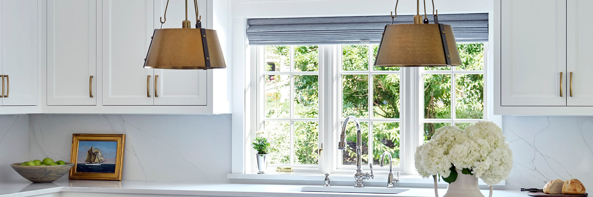 Bright white kitchen featuring Pella casement window above sink with brass pendant lights and marble countertops