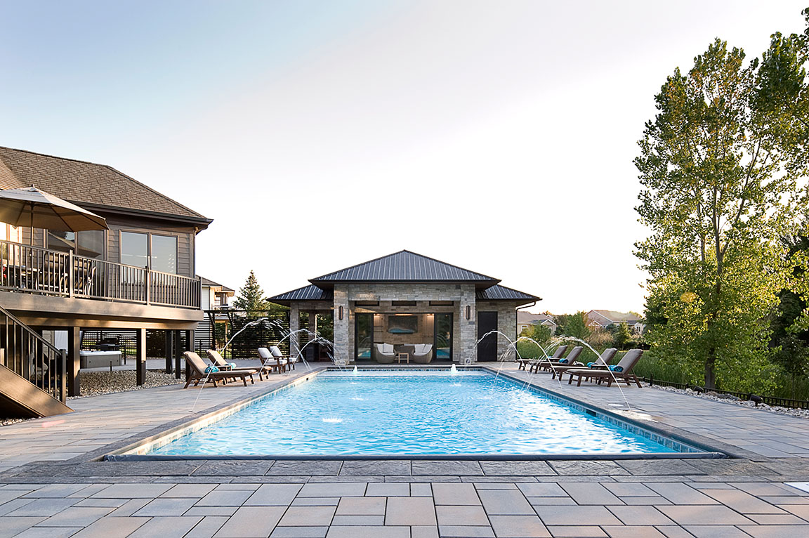 A modern pool house with large windows, featuring a stone patio and a dining area with wooden chairs.