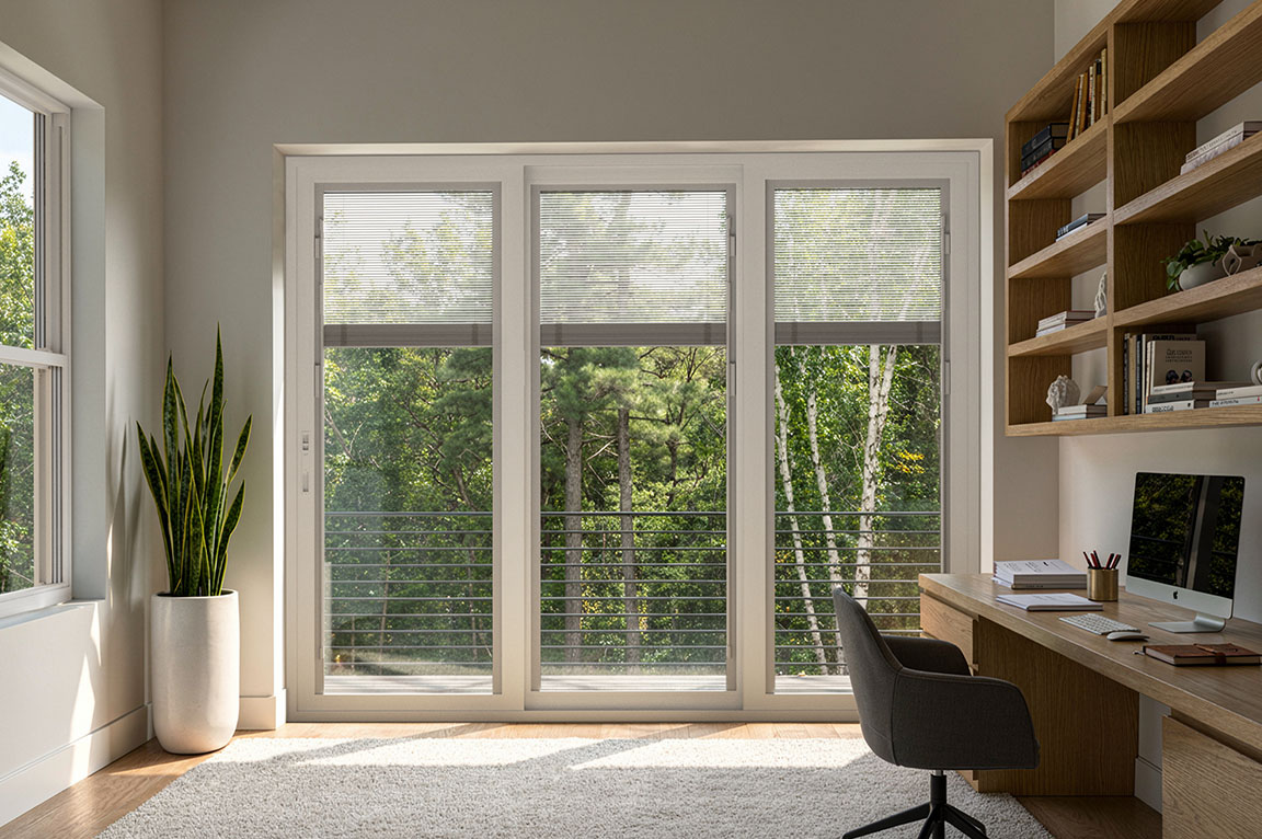 Home office with sliding glass doors and integrated blinds facing a wooded landscape.