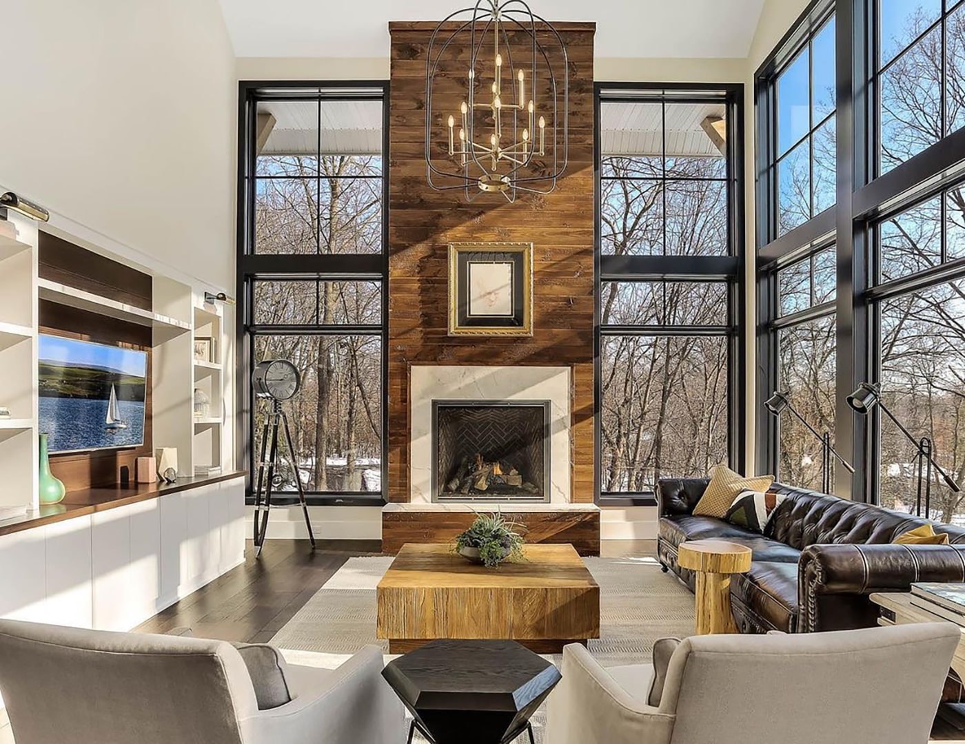 A living room with a high ceiling has floor to ceiling windows and a tall wood fireplace, topped with a grandiose chandelier.