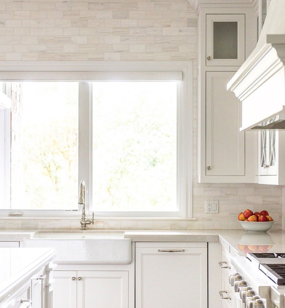 A traditional kitchen with white cabinets and backsplash features casement windows above the sink.