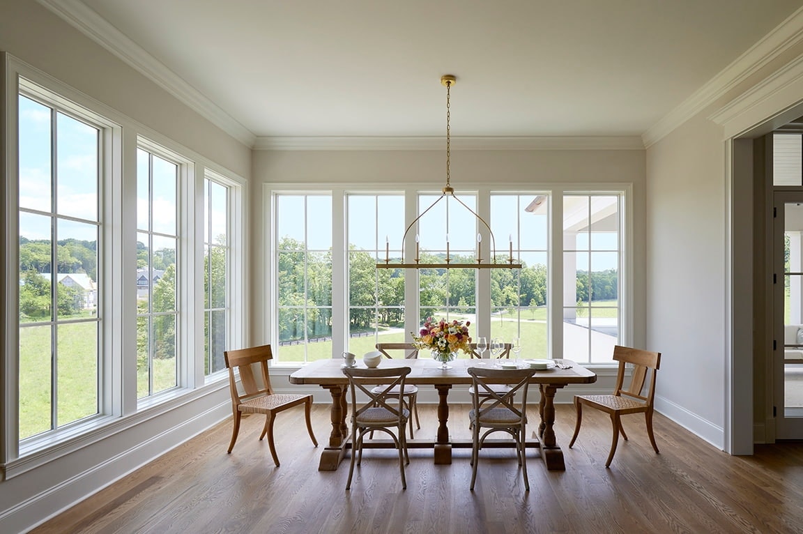 A bright dining room with large windows, a wooden table, and chairs, featuring elegant interior window trim.
