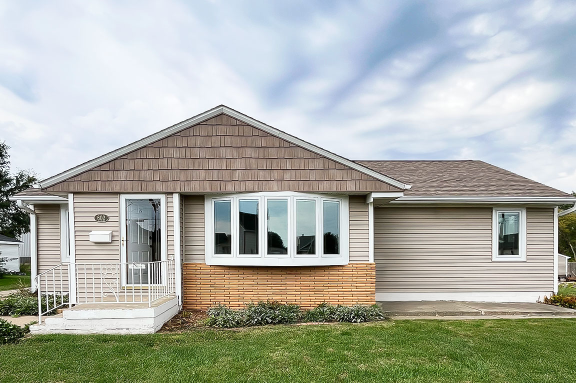 Single-story home exterior with tan vinyl siding, brick accent, and a large white bay window on the front facade.