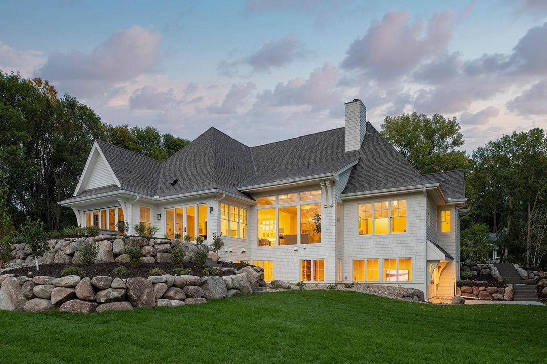 The backside of a farmhouse showcases many white windows illuminated at dusk.