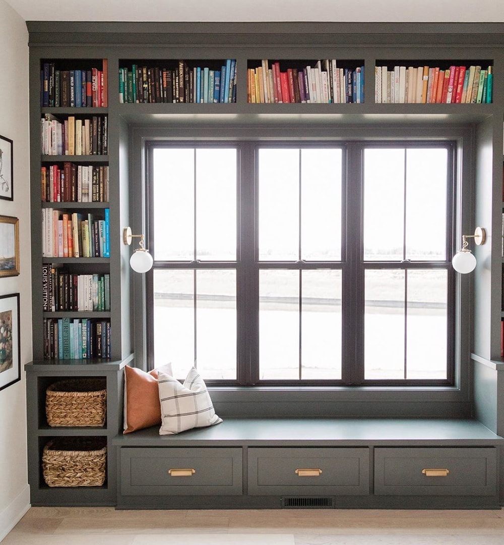 At the center of gray built-in bookshelves and a window seat are three double-hung windows with simple grilles.
