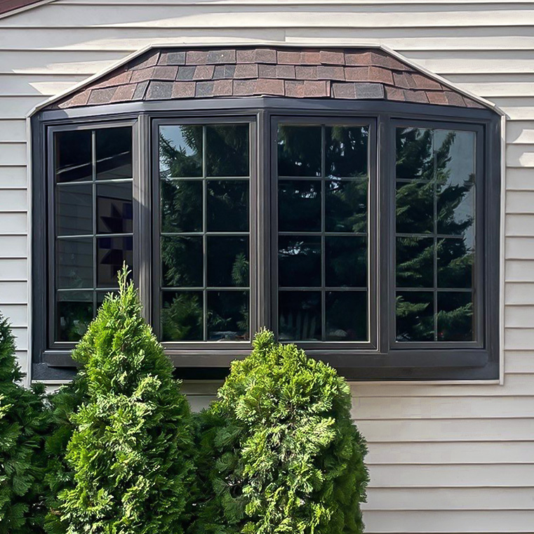 Bow window with brown frame on tan siding, surrounded by green shrubs.