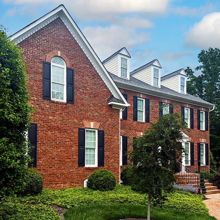 A large, elegant brick colonial house featuring black shutters, white trim, and multiple dormer windows on the roof. The house is surrounded by well-maintained greenery and a clear blue sky with some clouds in the background.