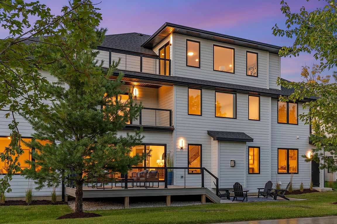 Rear exterior of modern home featuring expansive windows, upper balcony, and backyard deck at dusk.
