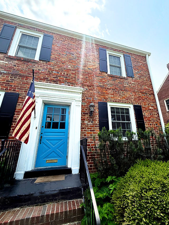 Close-up of the front entrance of a red brick Colonial house featuring a blue door with a white decorative frame, an American flag mounted to the left, black railings, and green shrubs lining the steps under bright sunlight.