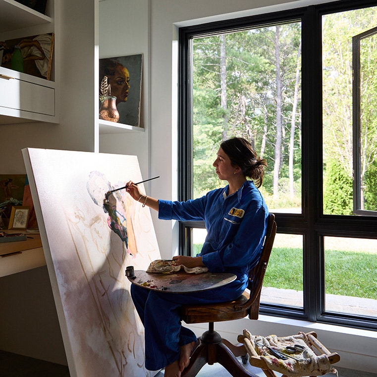 An artist painting on a canvas in a modern studio with a casement window, surrounded by greenery.