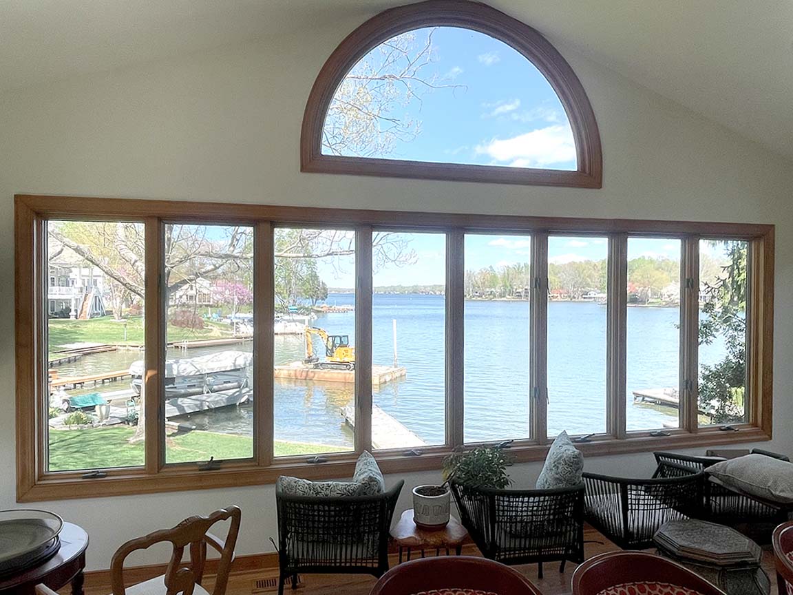 A living room featuring a large window with multiple panes and a semi-circular top section, providing a picturesque view of a lake, docks, and surrounding greenery. The room contains chairs and cushions arranged near the window.