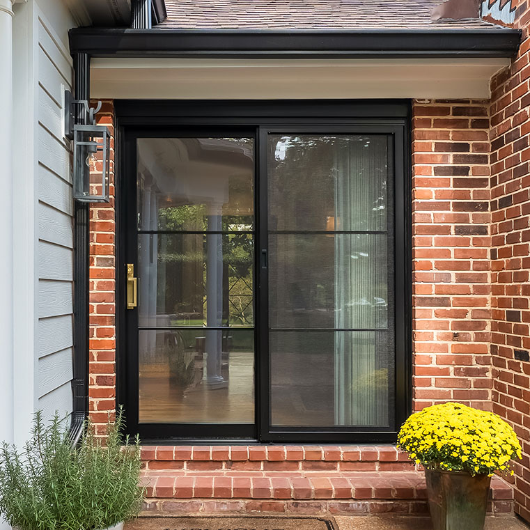 Black sliding patio door with full glass panels and brass handle on brick exterior.