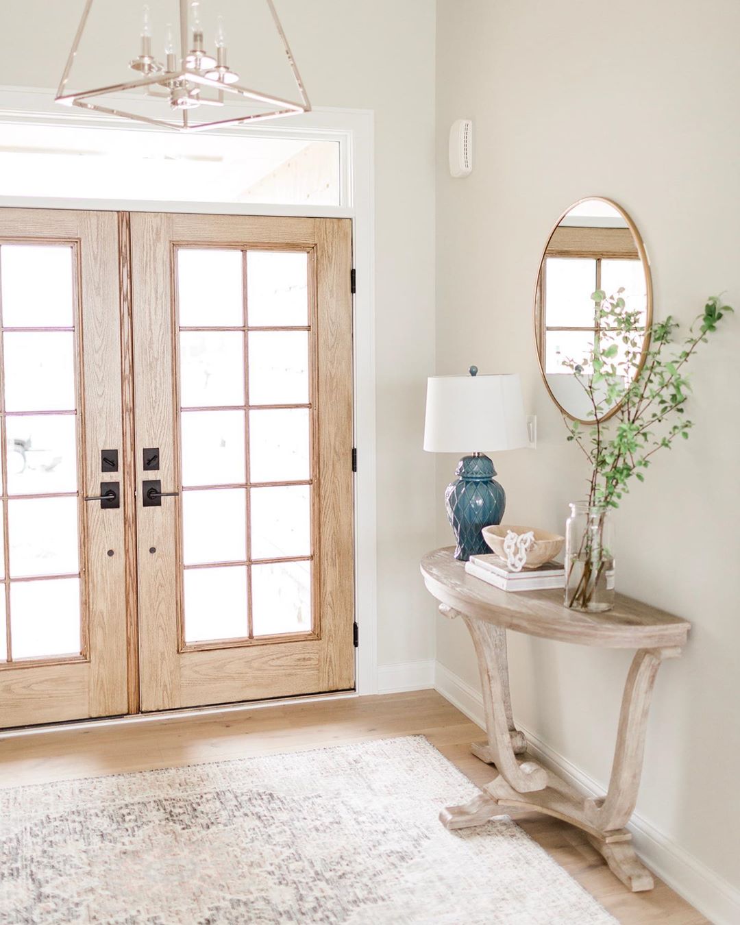 entryway with oak french patio doors and console table with blue lamp, round mirror, and greenery