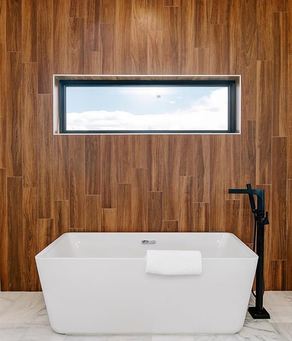 A stunning soaking tub sits in front of a wood-board wall, and a single wide awning window.