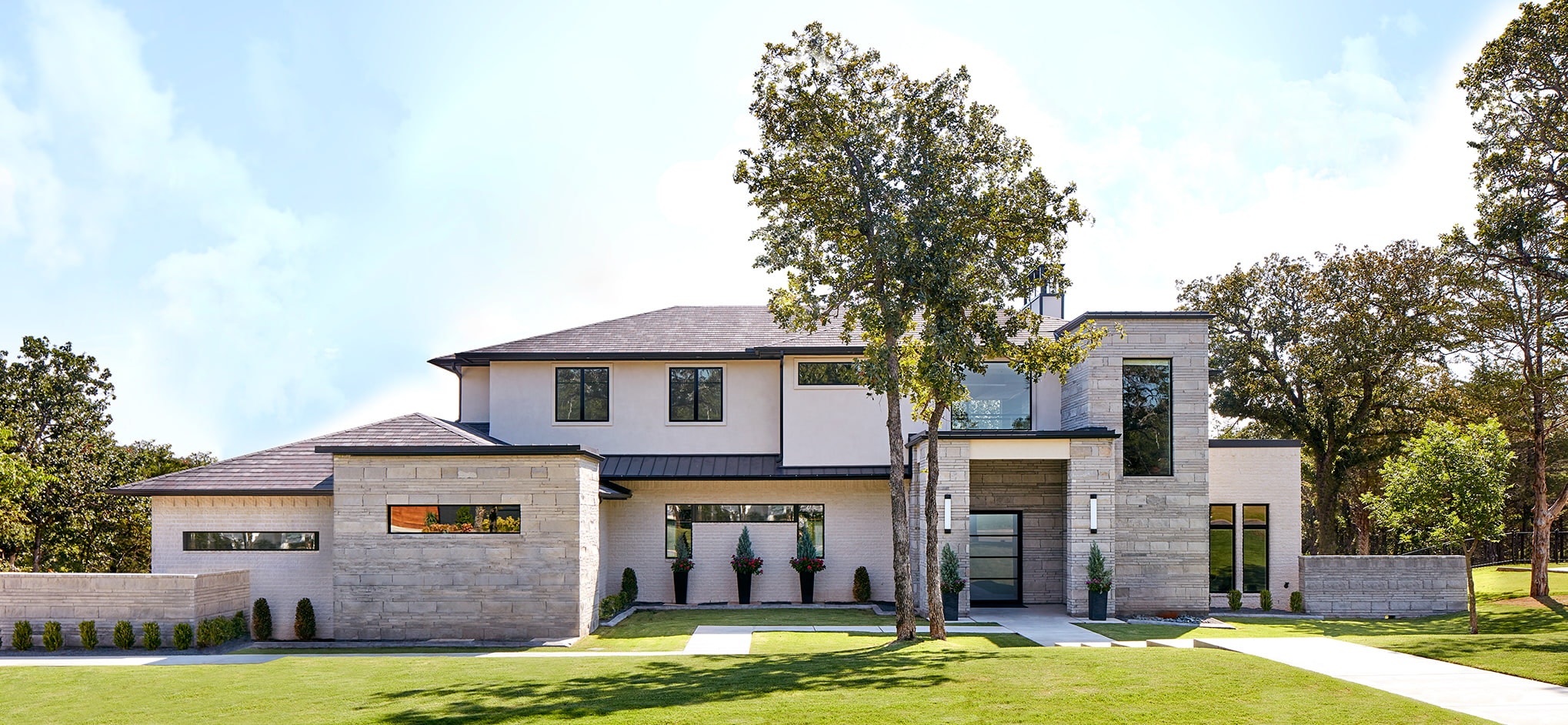 The exterior of a modern home in Oklahoma has stone, black fiberglass windows and beautiful landscaping.