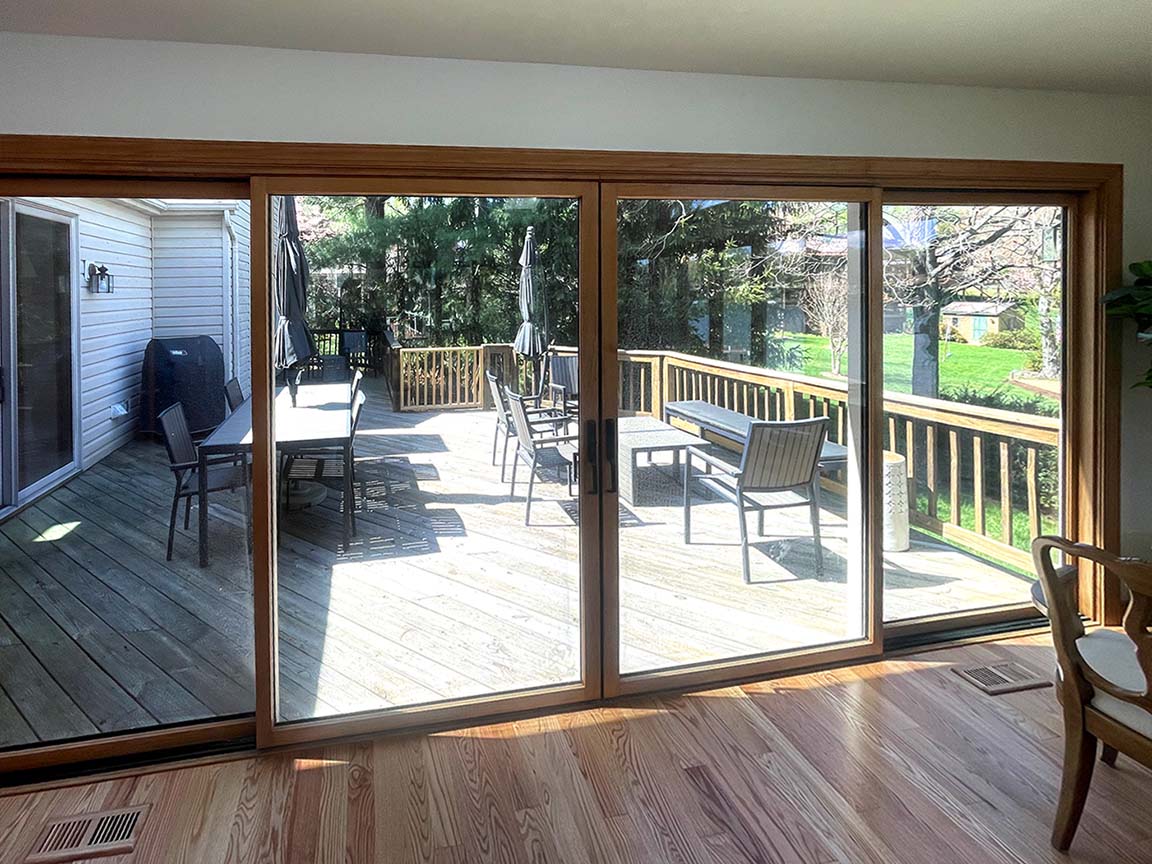 View of a spacious wooden deck with outdoor tables, chairs, and umbrellas seen through large sliding glass doors.
