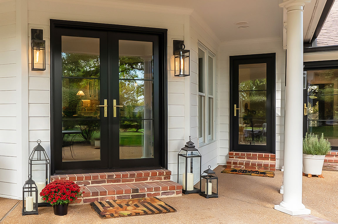 Black French doors with glass panels and brass handles on white siding exterior with lantern decor.