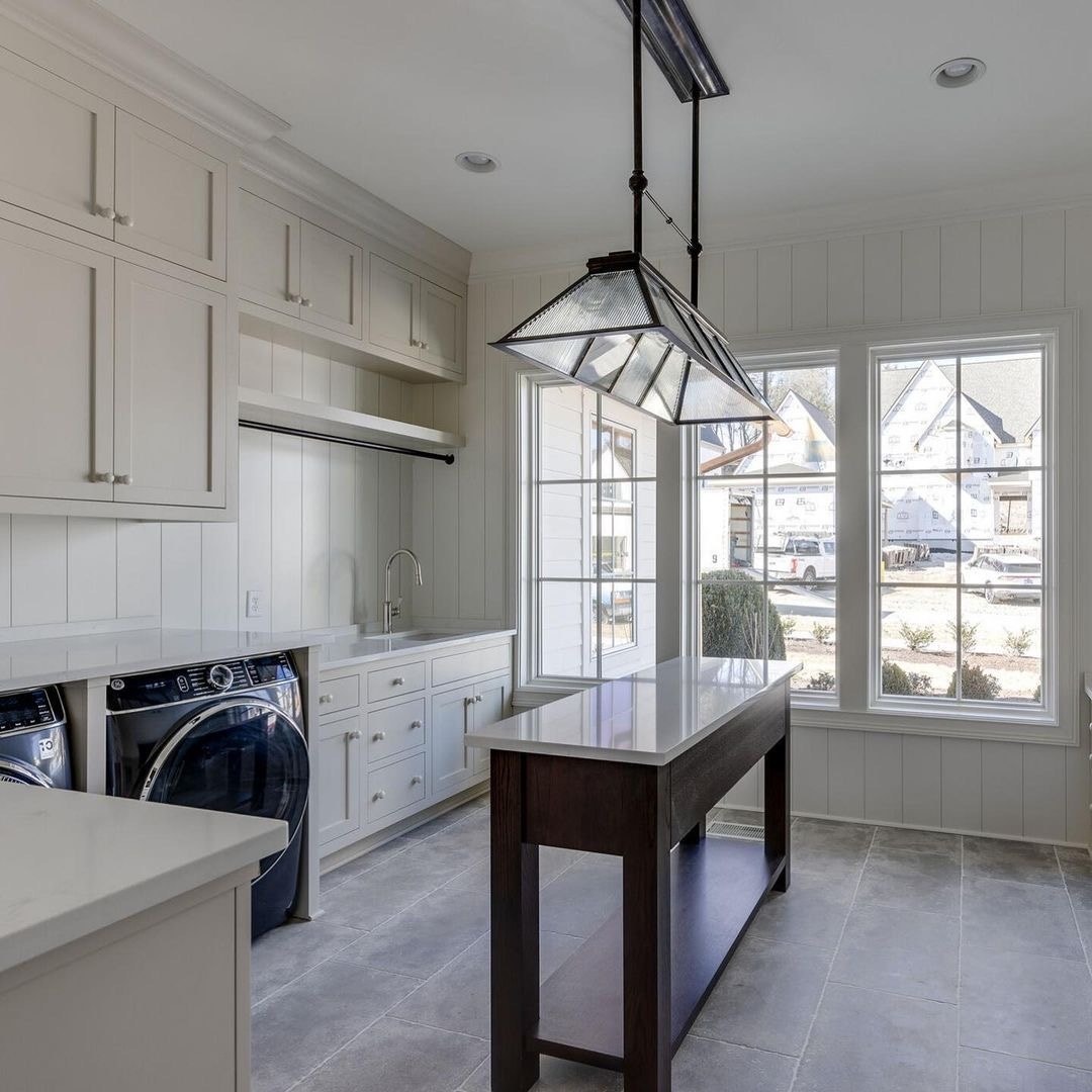 A cream-colored laundry room has a table in the middle with traditional windows behind it.