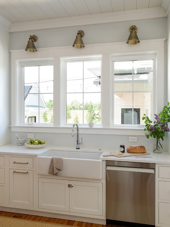 A modern kitchen with white cabinets, a farmhouse sink, and interior window trim, featuring natural light and greenery.