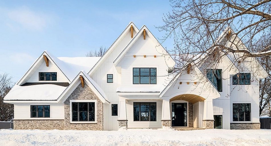 A large white farmhouse features black double-hung windows, wood corbles and warm-toned stonework
