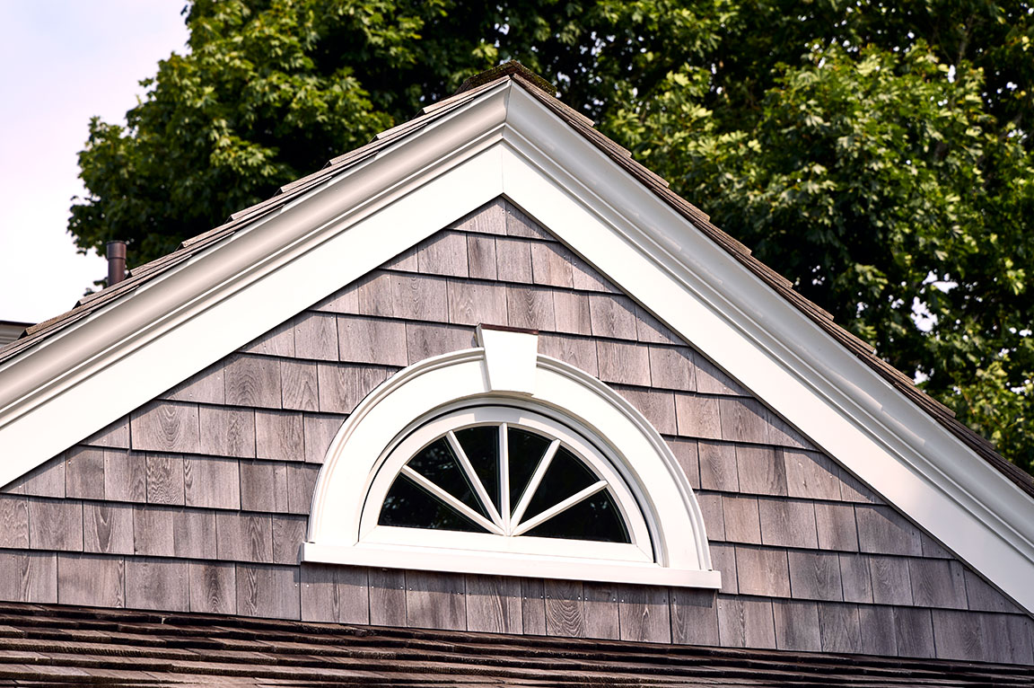 An architectural detail of a sun burst grille pattern window set in a gabled roof with wooden shingles.