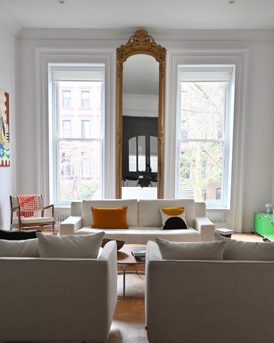 Two white double-hung windows are placed on either side of a gold mirror in a living room.
