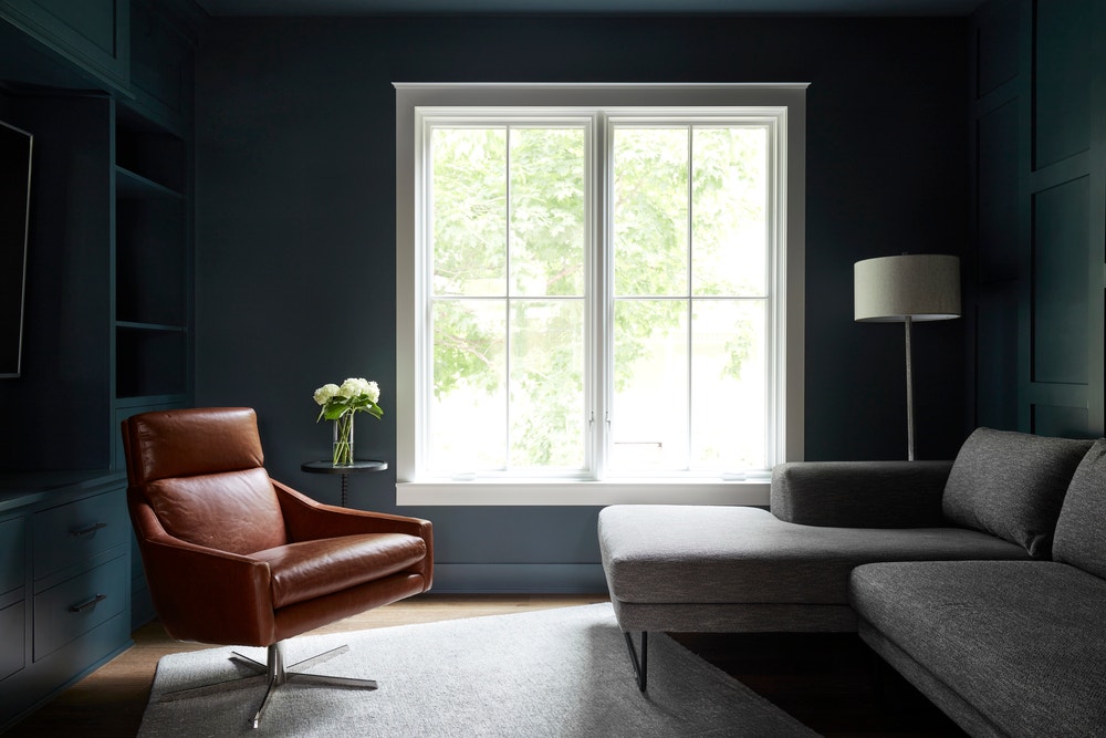 dark living room walls contrasts with two white casement windows and brown leather chair