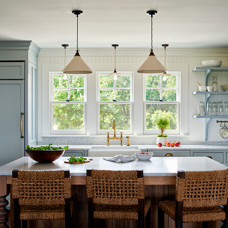 A modern kitchen with large windows allowing natural light, featuring bar stools and a bowl of fresh vegetables.
