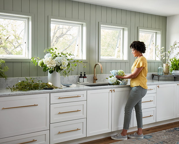 A woman wearing a yellow top is arranging fresh flowers at the sink which is underneath square awning windows