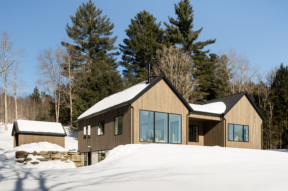 A modern house in a snowy landscape, featuring large fiberglass windows and surrounded by trees.