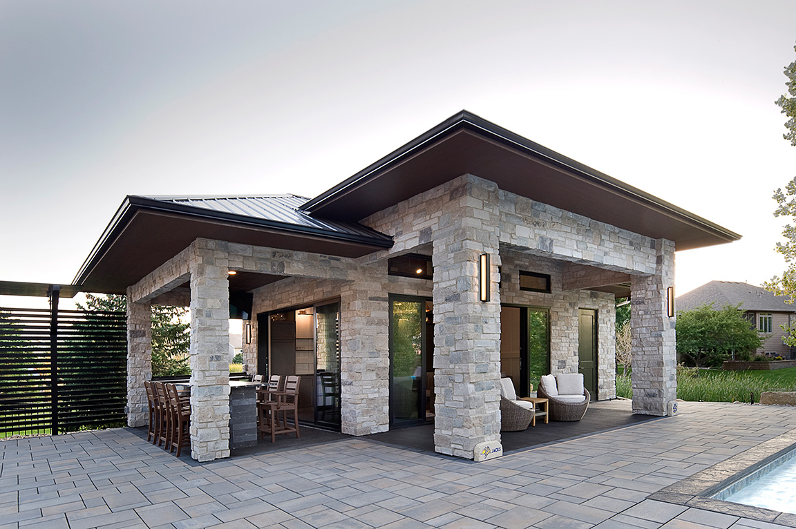  A modern stone pool house with large windows, featuring a patio and outdoor seating area.