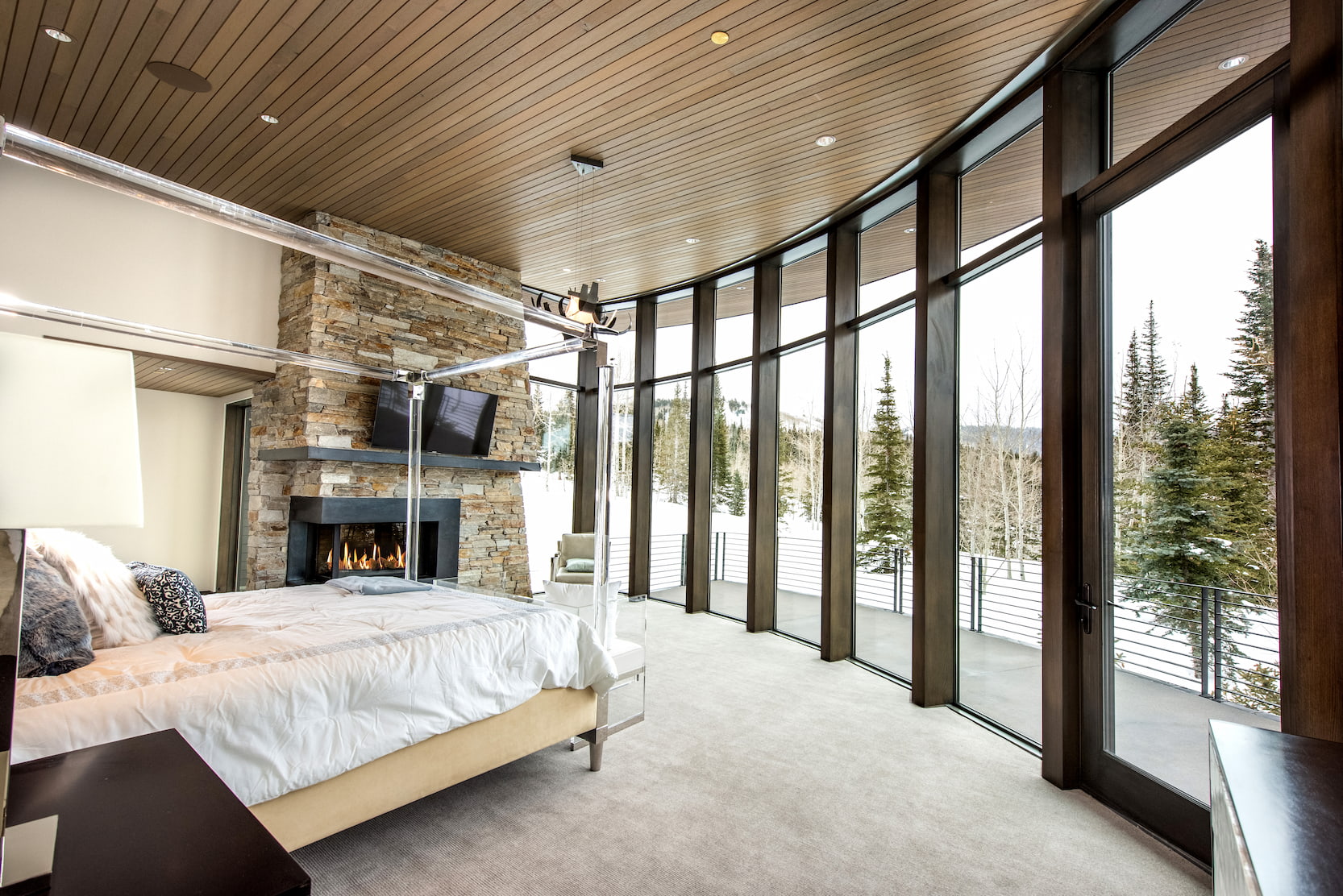 A bedroom with wood ceilings and a fireplace features a curved window wall with a hinged patio door.